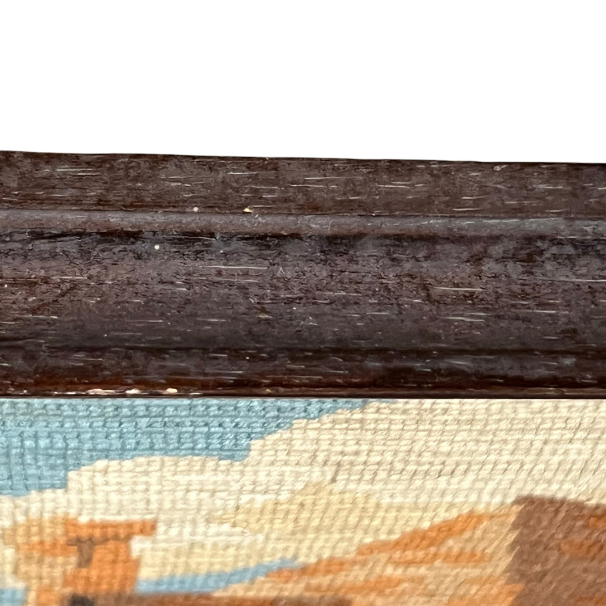 Close-up detail of a framed needlepoint canvas, showing the dark brown wooden frame top and the woven canvas texture with blue, beige, and orange colors.
