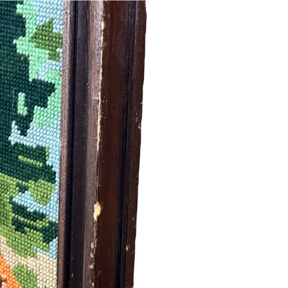Close-up of a needlepoint canvas showing a landscape design with green trees, blue sky, and orange ground, bordered by a dark brown wooden frame on a white background.