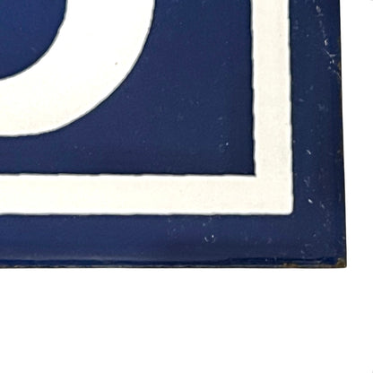 A close-up of a square decorative tile or sign corner with a dark navy blue background, white border, and part of a white curved shape visible.