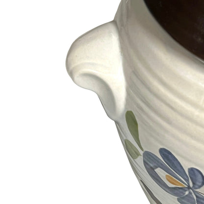 Close-up of a white ceramic pitcher with a curved handle, featuring a blue and green floral pattern and a dark brown interior, set against a plain white background.