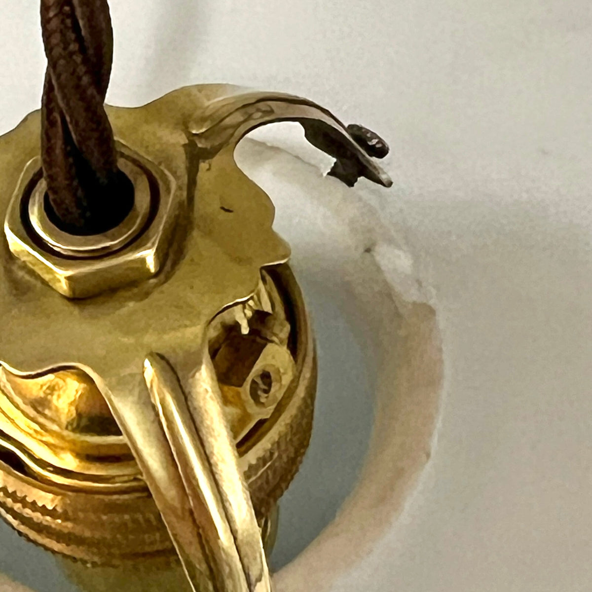 Close-up of a brass ceiling light fixture base with a dark brown fabric cord entering from the top, set against a white background.