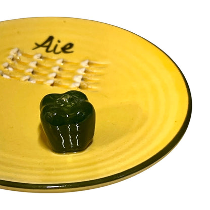 A yellow plate featuring a green pepper and the French word for garlic, “Ail,” designed for grating garlic and perfect for a French farmhouse-style kitchen, set against a white background.