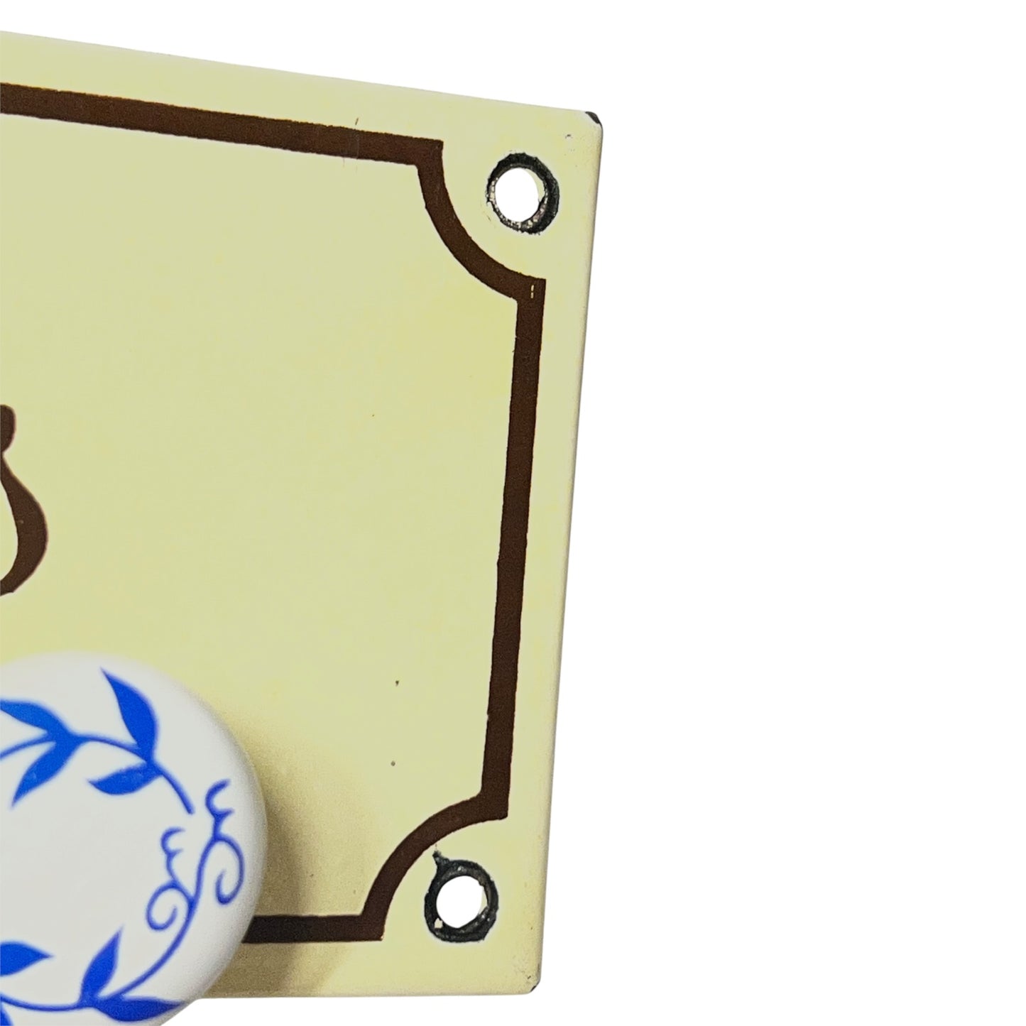 Close-up of a cream-colored decorative sign or plaque with dark brown border lines and mounting holes, partially overlaid by a round white knob with blue floral scrollwork, on a white background.