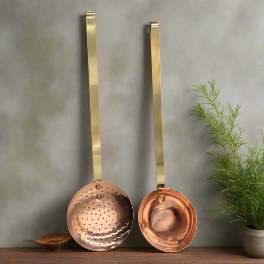 Set of 2 French copper utensils with long brass handles, includes a straining spoon and a ladle on a grey background beside a plant