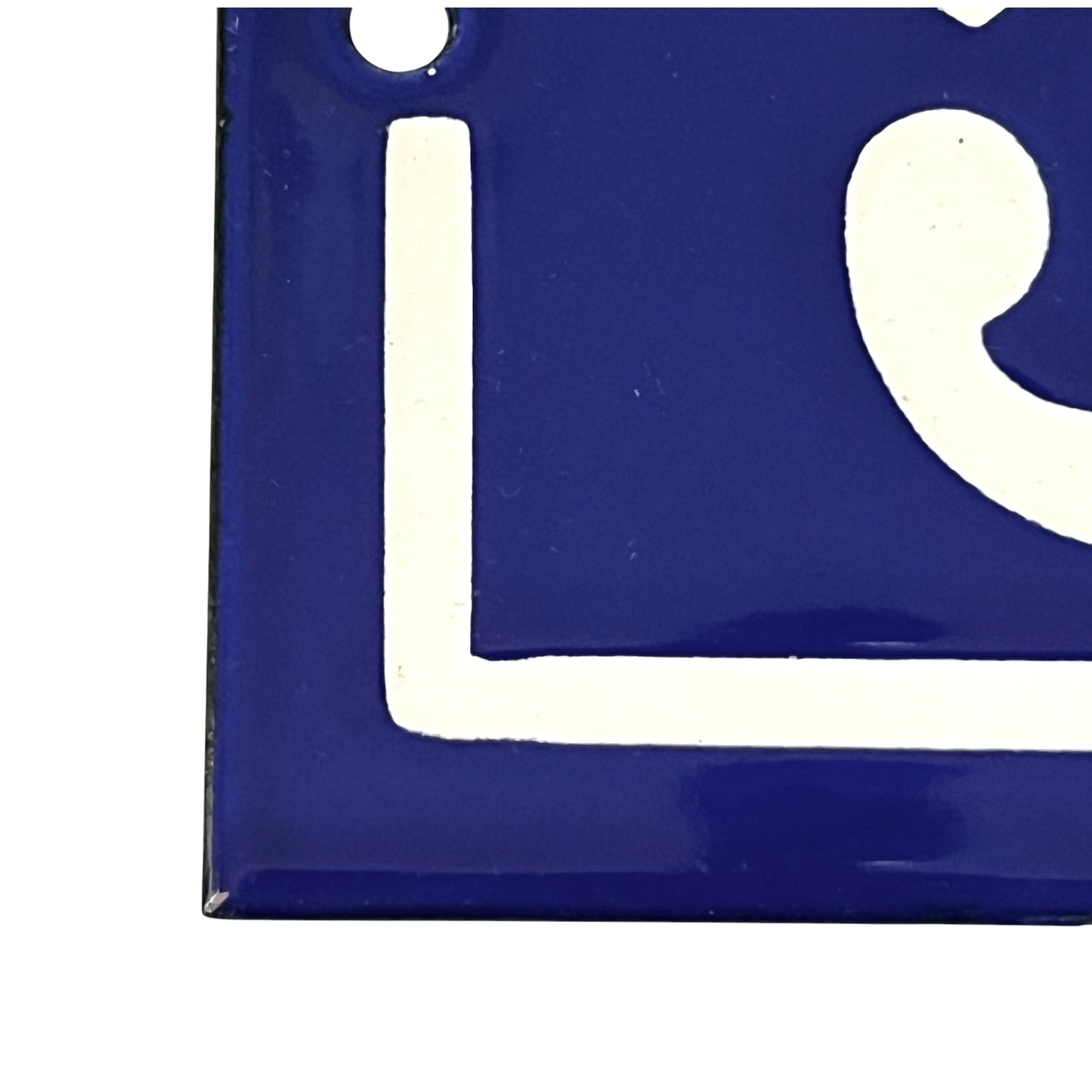 A close-up of a glossy blue square tile or sign with a white painted letter or symbol, shown against a white background.