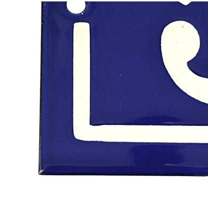 A close-up of a glossy blue square tile or sign with a white painted letter or symbol, shown against a white background.