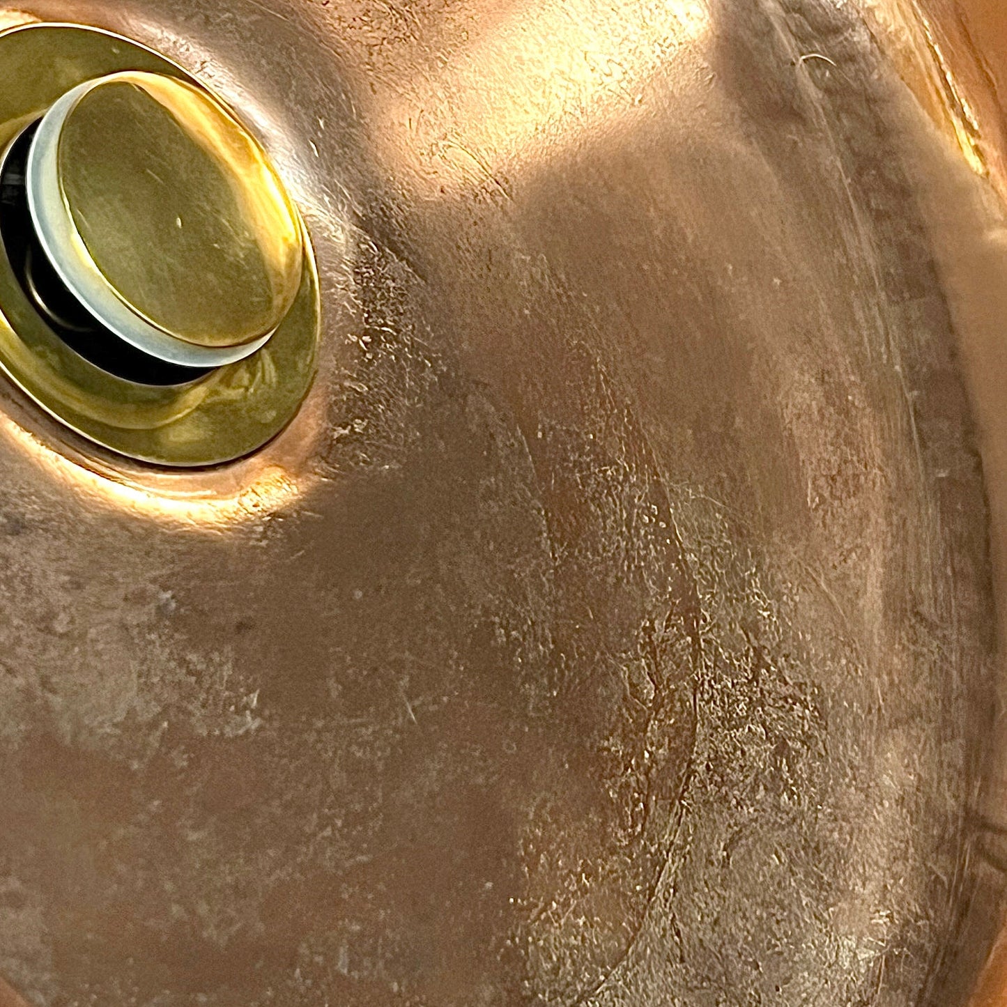Close-up of a brushed metallic sink basin with a round drain and pop-up assembly, showing a warm copper-bronze finish with reflective highlights.