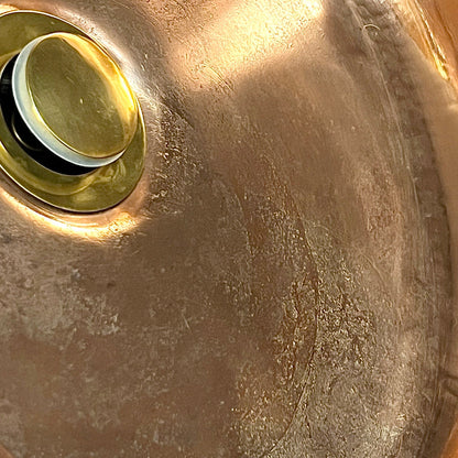 Close-up of a brushed metallic sink basin with a round drain and pop-up assembly, showing a warm copper-bronze finish with reflective highlights.