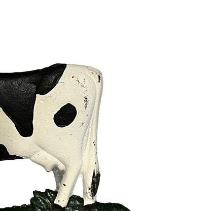 A close-up of a black and white cow figurine standing on a dark green textured base against a plain white background.