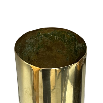 A close-up of a polished brass cylindrical container or vase, showing a reflective metallic gold exterior and a dark green patina inside, set against a plain white background.