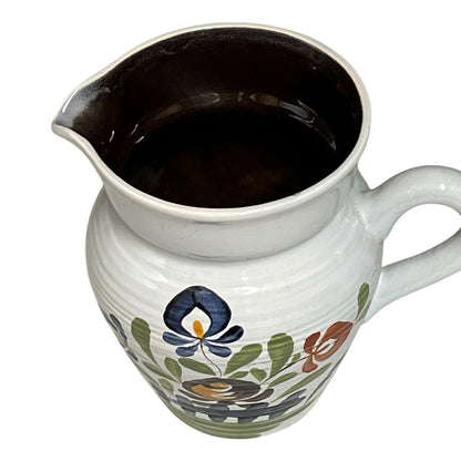 A white ceramic pitcher with a dark brown interior and a hand-painted floral pattern in blue, green, and orange on the front, shown against a plain white background.