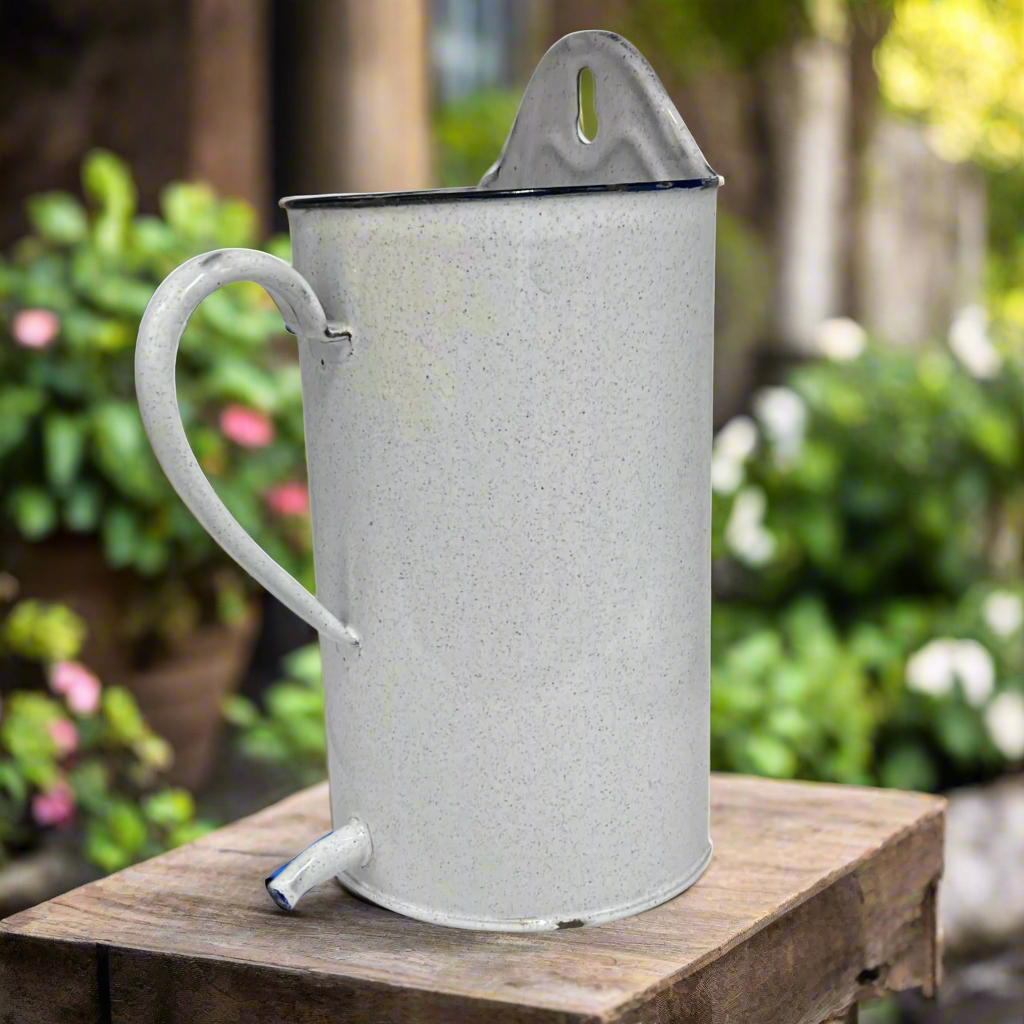 White speckled enamel jug or planter with a spout on a table in a garden with a blurred background of flowers for sale by All Things French Store