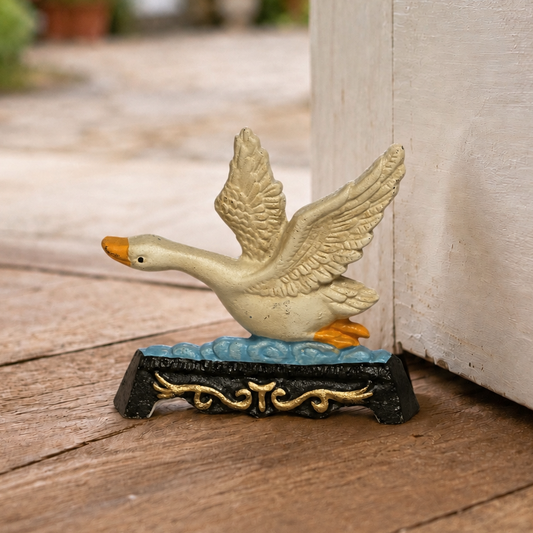 A decorative goose doorstop with wings raised, standing on a blue wave-like base with ornate gold detailing, placed on a wooden surface beside a white door in an outdoor setting.