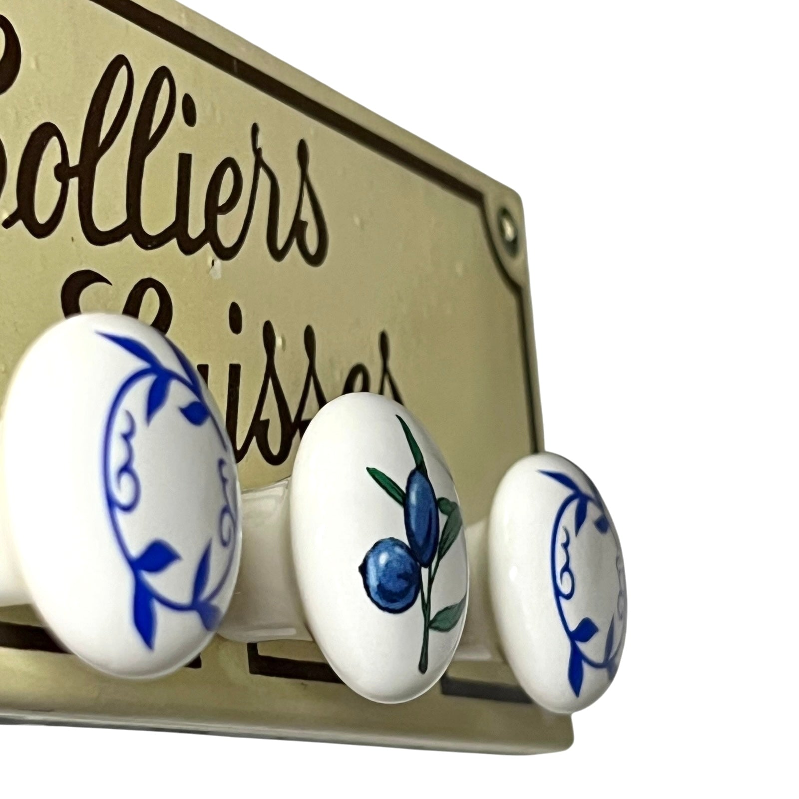 Three round white ceramic cabinet knobs with blue and green floral and berry designs, mounted on a beige wooden sign with the words 'Colliers Laisses' partially visible.