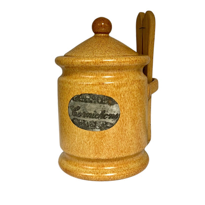 A rustic ceramic cornichon jar with a lid and  tongues, featuring a grey oval label on the front that reads 'Carnichons'.