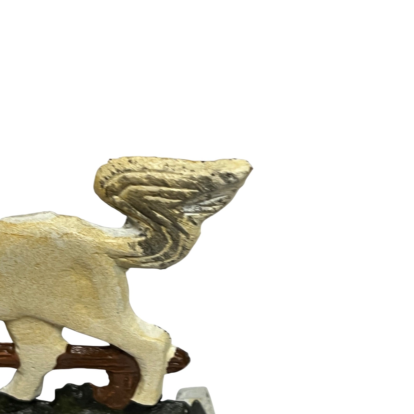 A close-up of a small winged animal figurine, likely a griffin or cherub, shown from the side on a white background. The figurine has a textured, weathered finish in light beige and gray tones, mounted on a dark brown base.