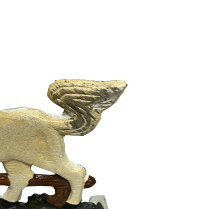 A close-up of a small winged animal figurine, likely a griffin or cherub, shown from the side on a white background. The figurine has a textured, weathered finish in light beige and gray tones, mounted on a dark brown base.
