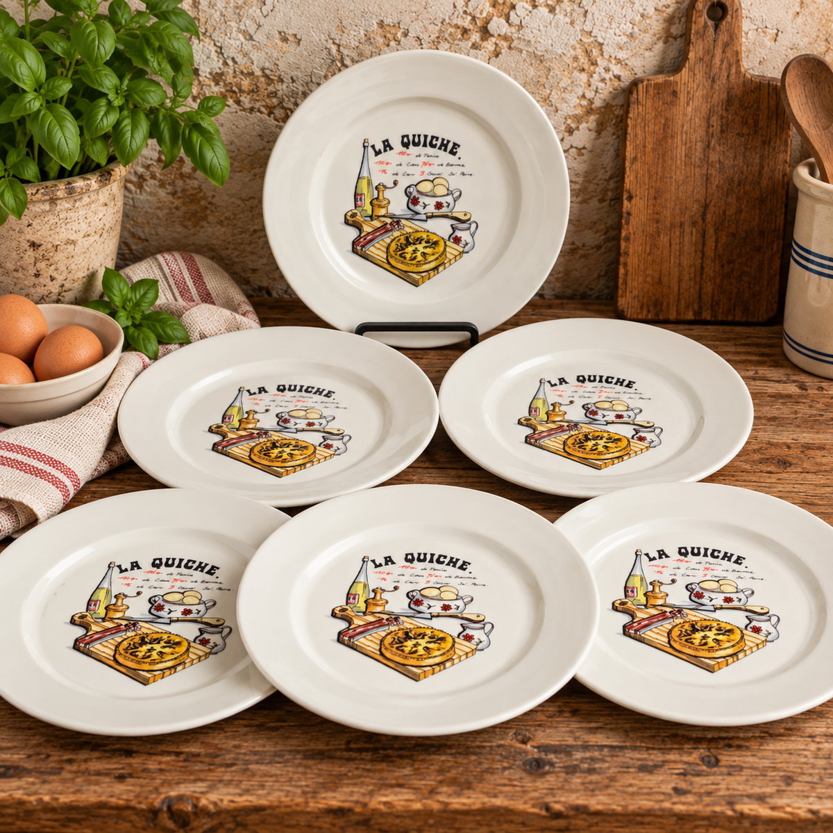 Set of six vintage French ceramic La Quiche recipe plates, each measuring 20cm in diameter, displayed in a rustic farmhouse kitchen. One white plate stands upright on a display stand, with five matching plates arranged naturally on a wooden table. Each features colourful illustrations of quiche, eggs and traditional French cooking ingredients with French recipe lettering. Styled against a textured stone wall with fresh herbs, eggs, striped linen and vintage wooden kitchen accessories.