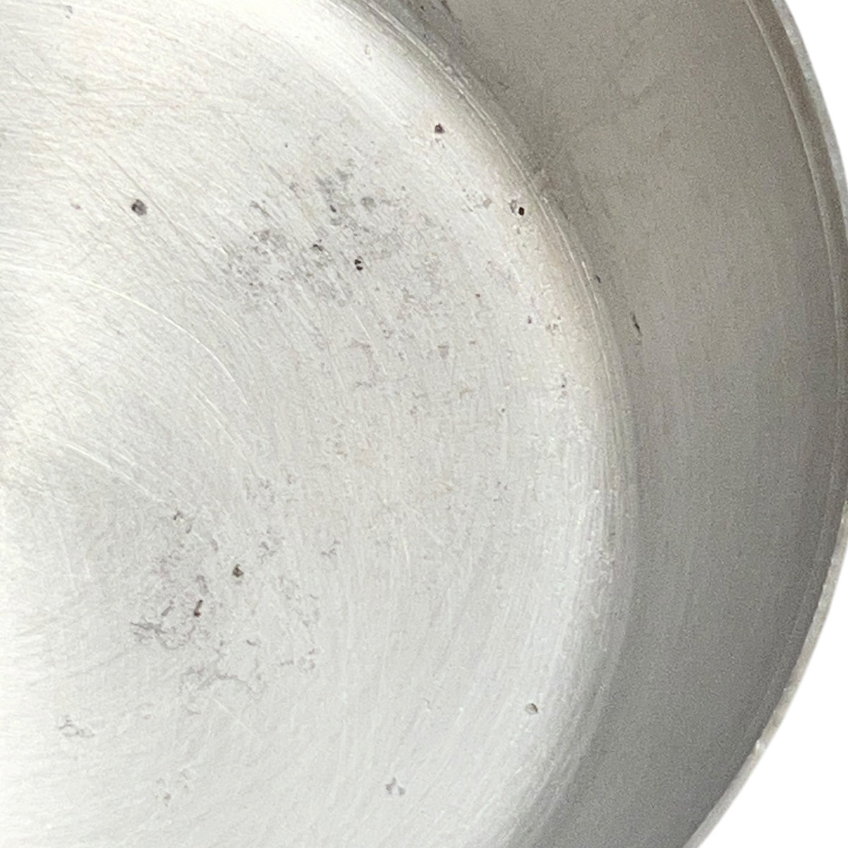 Close-up of the inside of a brushed metal pan or pot showing a silver aluminum surface with some minor scuffs and dark specks, against a white background.