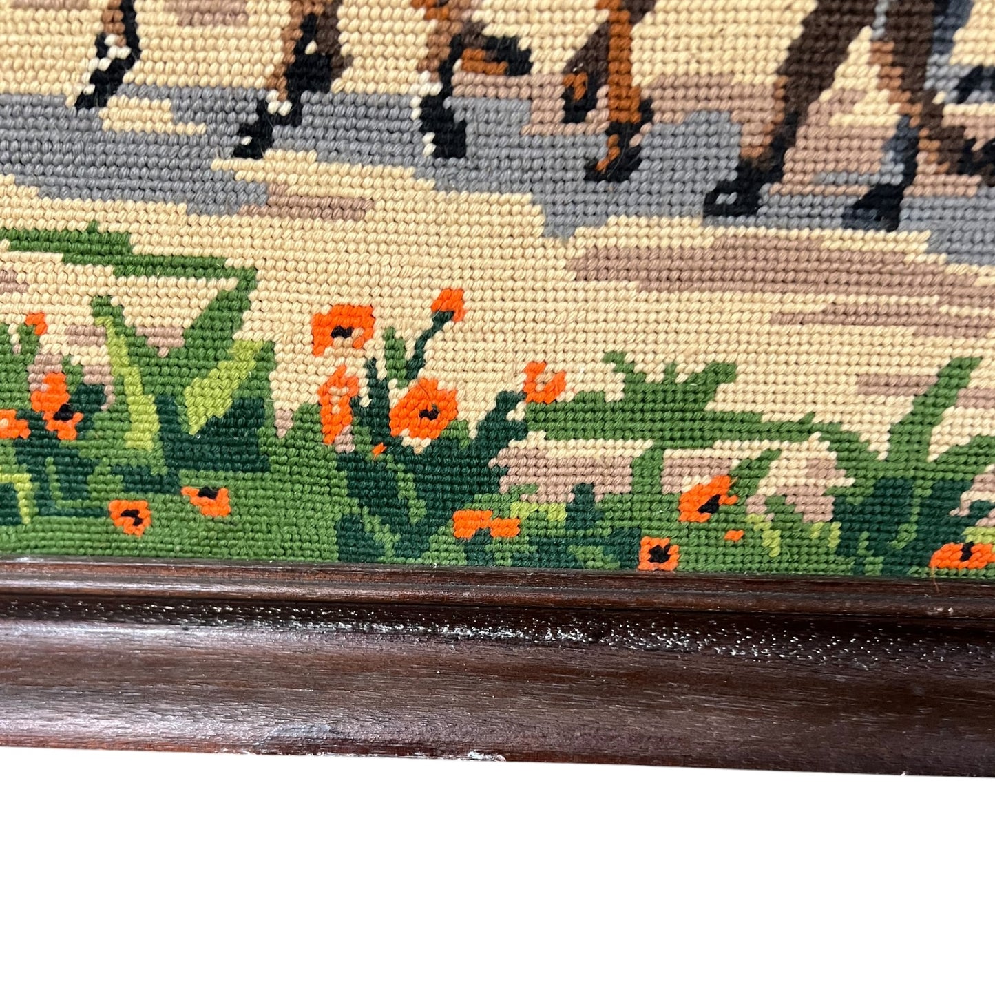 Close-up of a framed needlepoint or embroidered wall art showing horse hooves along the top, green foliage and orange flowers along the bottom, and a dark wooden frame.