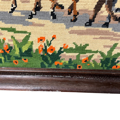 Close-up of a framed needlepoint or embroidered wall art showing horse hooves along the top, green foliage and orange flowers along the bottom, and a dark wooden frame.