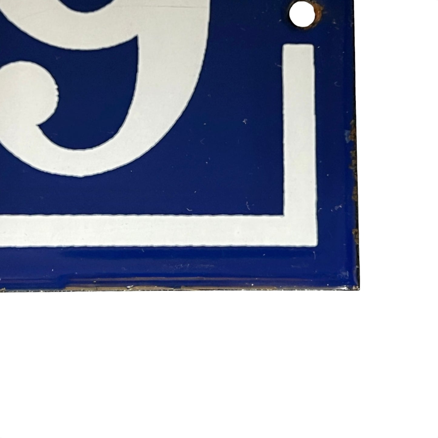 Close-up of a blue square sign or plaque with a white stylized number and corner border, showing slight wear and a small hole near the top right, against a white background.