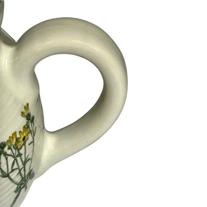 Close-up of a white ceramic pitcher with a curved handle and a delicate yellow and green floral decoration on the side, set against a plain white background.