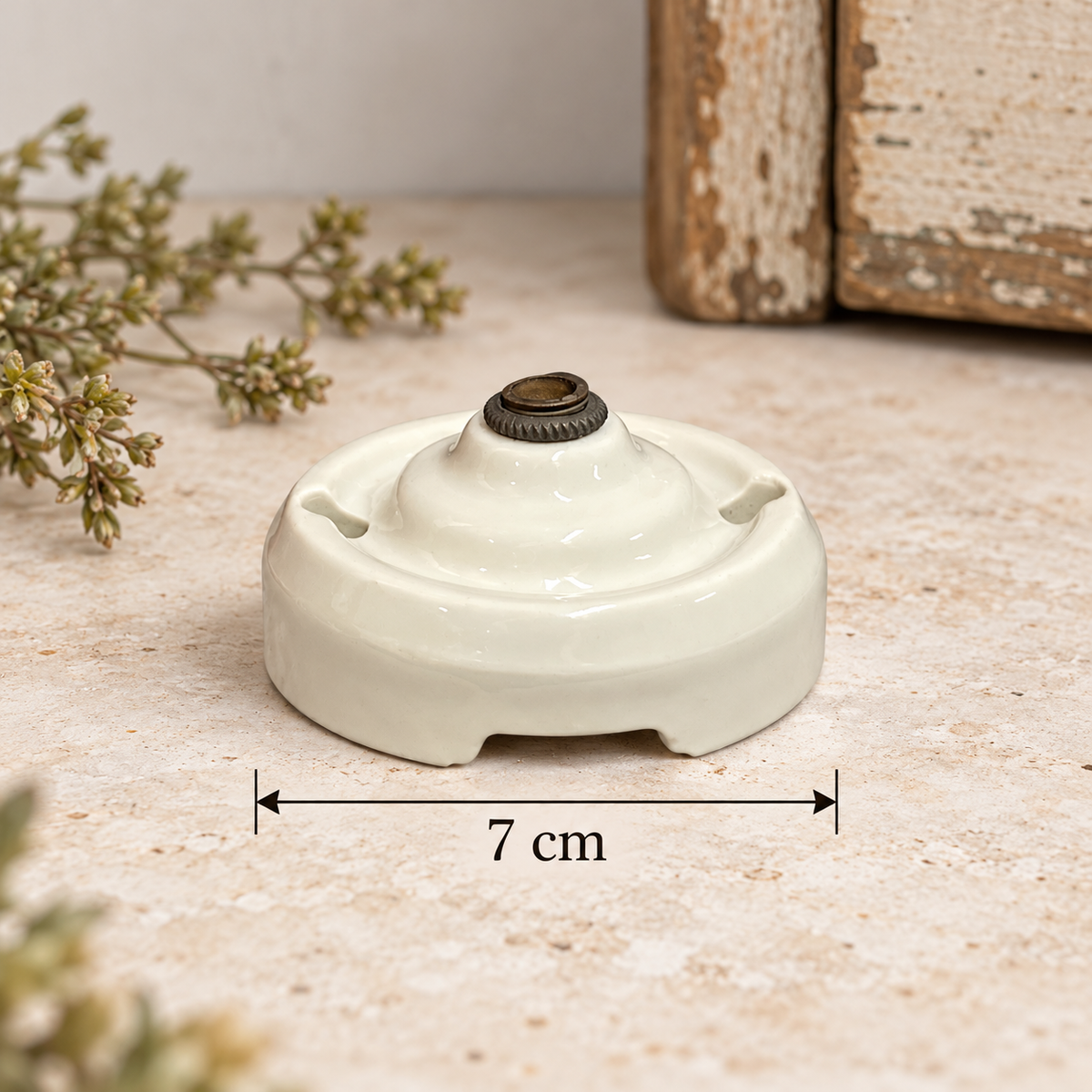 A small round ceramic object with a glossy off-white finish and a dark metal top, shown on a textured beige surface with a dimension marker indicating 7 cm width. Decorative dried flowers and rustic wood appear in the background.