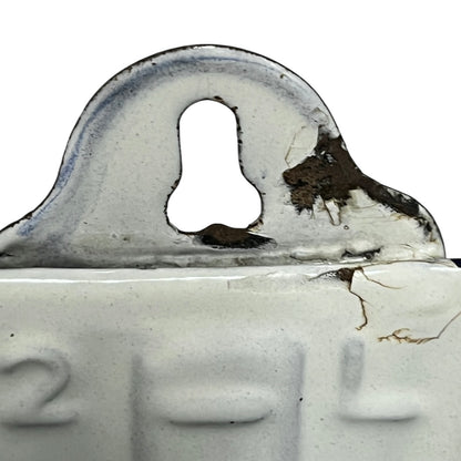 Close-up of a vintage white enamel wall container showing embossed numbers and a weathered metal top with rust and peeling paint against a white background.