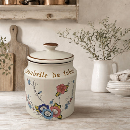 Decorative kitchen canister with floral designs, a lid and text Poubelle de Table on a wooden surface.