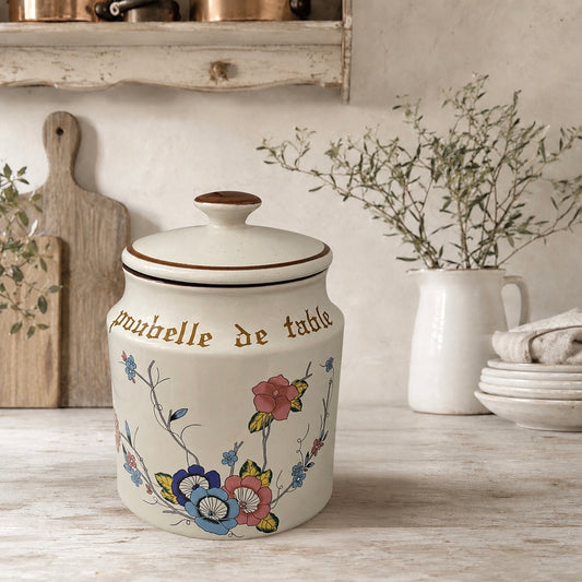 Decorative kitchen canister with floral designs, a lid and text Poubelle de Table on a wooden surface.