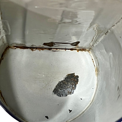 A close-up view inside a white enamel pot or container, showing a dark, textured metallic piece of scale or rust accumulation on the bottom floor, with brown rust stains along the rim.