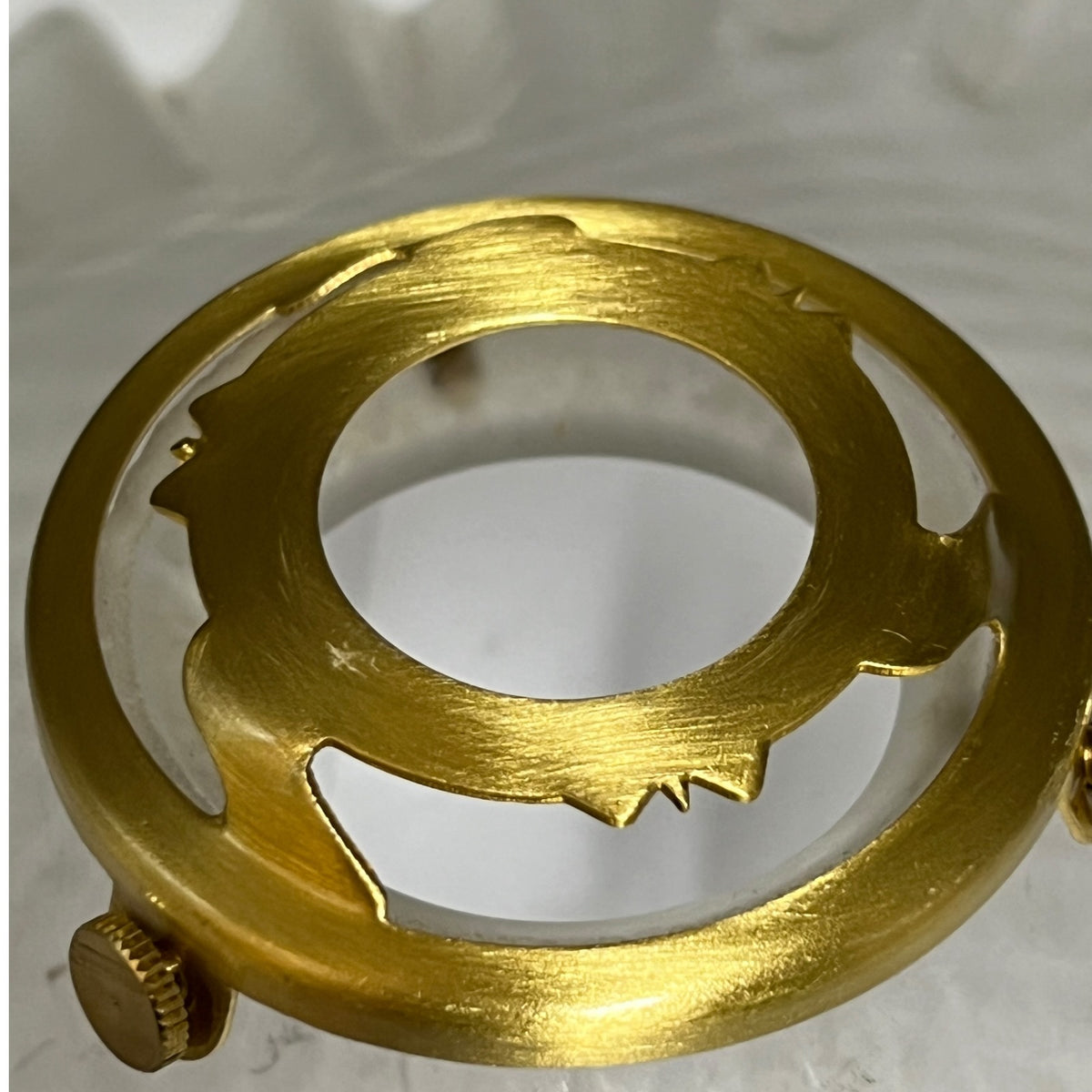 A close-up of a circular brass shade collar with cutout details and a crown knob, photographed against a light neutral background.