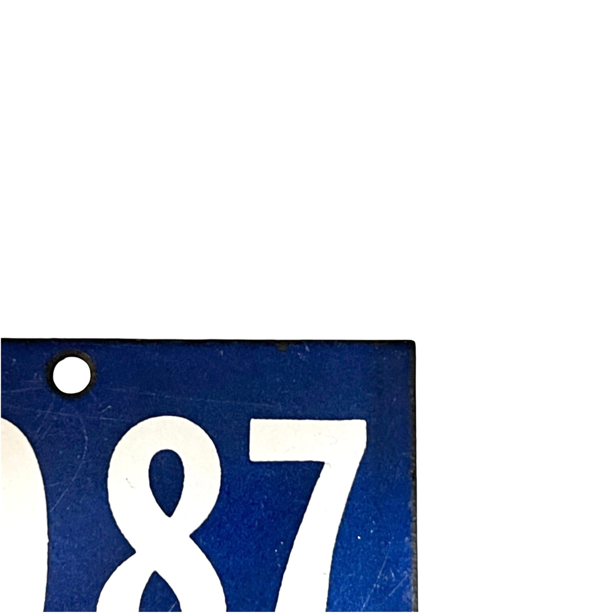 A close-up corner of a blue rectangular sign or plaque with large white numerals '87' and a small round hole near the top left, shown on a white background.
