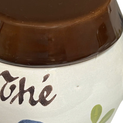 Close-up of a ceramic container with a glossy dark brown lid and an off-white body featuring dark brown script lettering and a small green leaf motif.