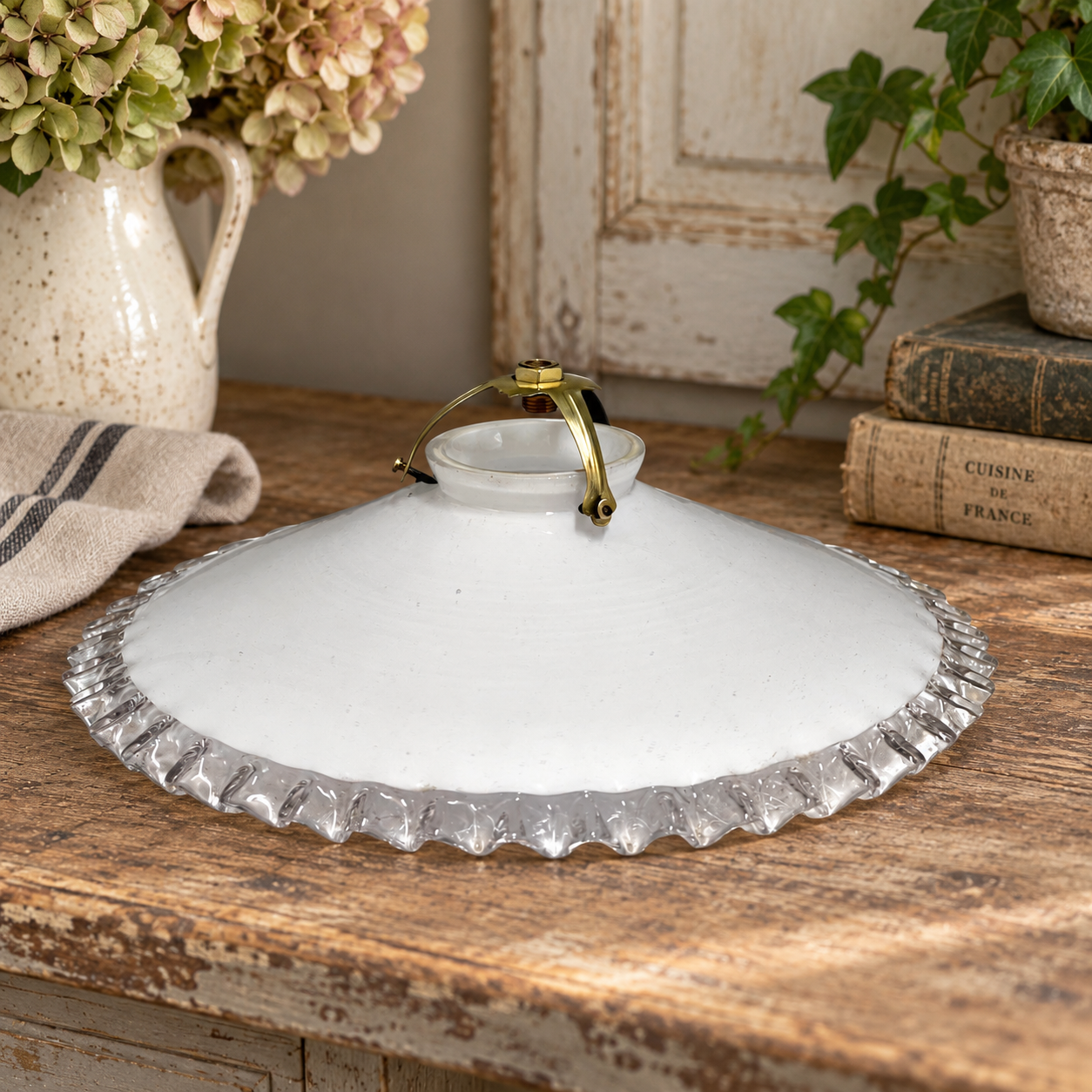 A white glass lampshade with a scalloped clear glass edge and a small gold-toned metal handle, displayed on a rustic wooden table with flowers, books, and greenery in the background.