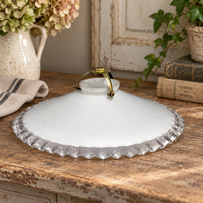 A white glass lampshade with a scalloped clear glass edge and a small gold-toned metal handle, displayed on a rustic wooden table with flowers, books, and greenery in the background.