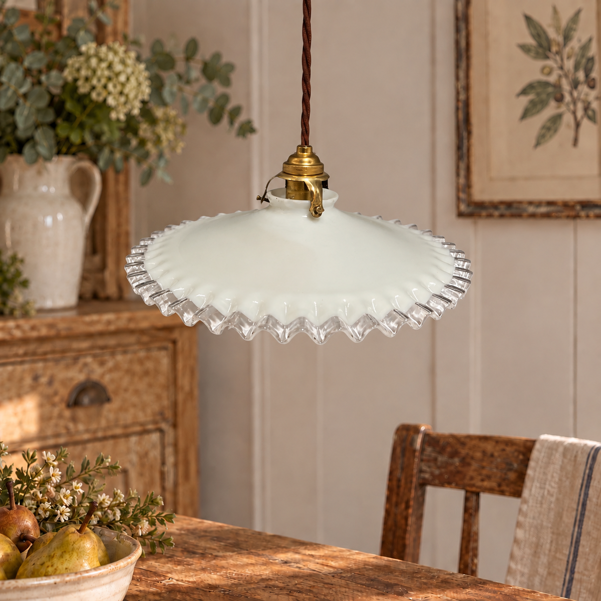 A vintage-style pendant light with a white ribbed glass shade and a brass fixture, hanging over a rustic wooden dining table in a warm, decorated room.
