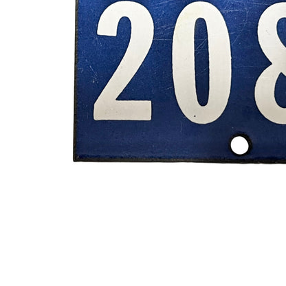 A close-up of a blue rectangular sign or plaque with large white numerals '208' and a small round hole near the bottom right, shown against a white background.