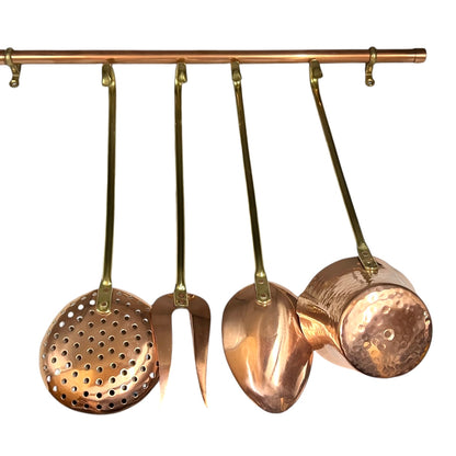 A set of hanging copper kitchen utensils on a brass rod, including a perforated skimmer, a spatula, a turner, and a small hammered pot, shown against a plain white background.
