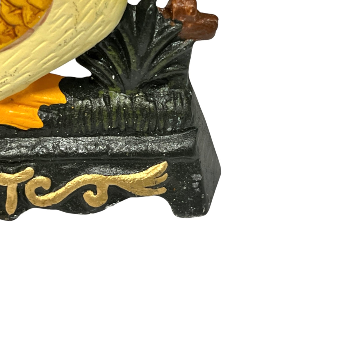 Close-up of a decorative figurine showing part of a yellow bird, dark green foliage, orange accents, and gold lettering on a black base against a white background.