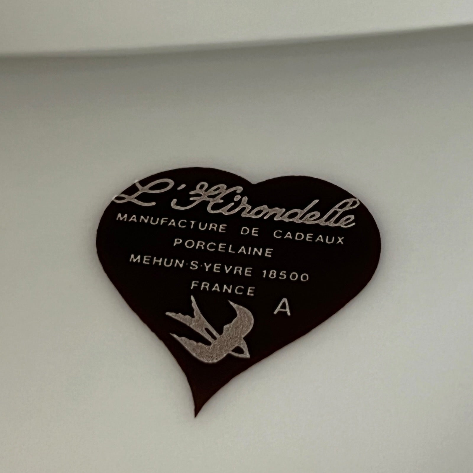 A heart-shaped dark brown plaque with white French text reading 'L'Hirondelle Manufacture de Cadeaux Porcelaine' and a silver bird emblem, photographed against a light neutral background.