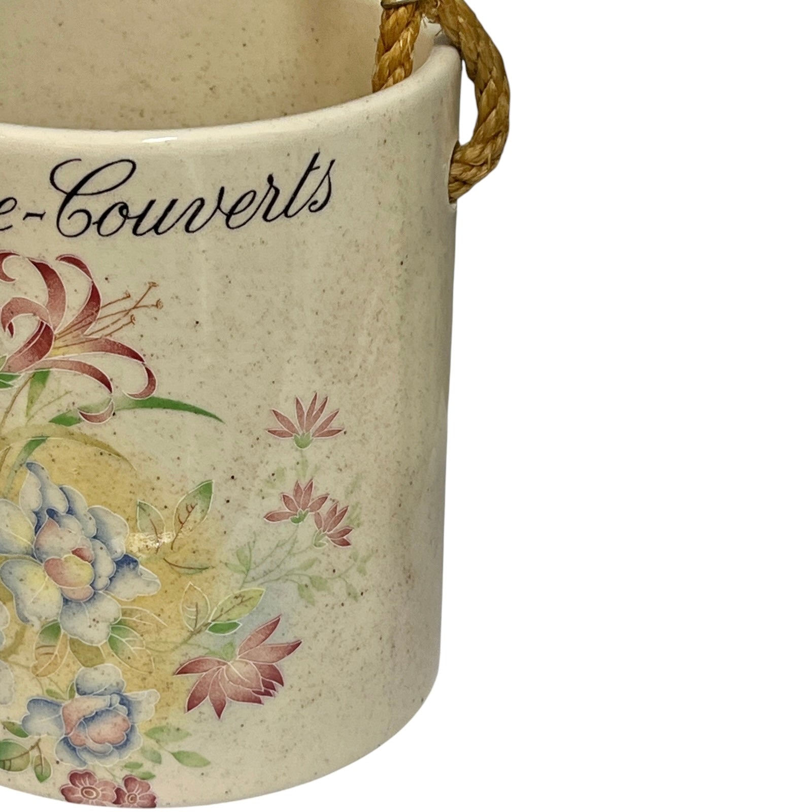 Close-up of a ceramic French crock pot or canister with a beige speckled glaze, featuring a floral print and the script text 'Couverlès', with a braided rope handle visible at the rim.