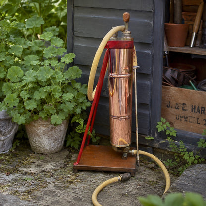 French vintage copper garden sprayer with a yellow hose and nozzle, standing upright on a wooden mount with a red-painted metal bracket. A wooden handle for pumping sits at the top, making it ideal for a vintage garden, all set against a rustic garden baclground