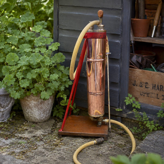 French vintage copper garden sprayer with a yellow hose and nozzle, standing upright on a wooden mount with a red-painted metal bracket. A wooden handle for pumping sits at the top, making it ideal for a vintage garden, all set against a rustic garden baclground