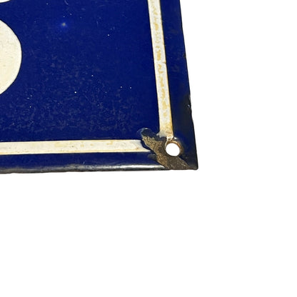 A close-up of a vintage blue enamel sign corner with a white painted border and a metal grommet, shown against a plain white background.