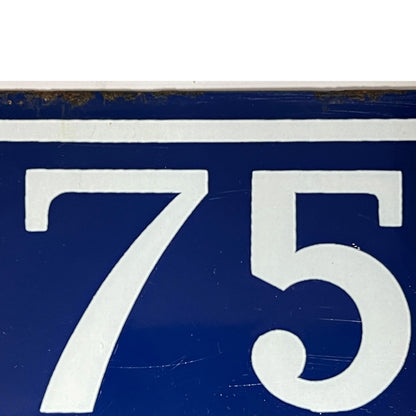 A close-up of a blue house number sign showing the number 75 in white, with a white horizontal stripe above the numbers and a small amount of weathered wear near the top edge.