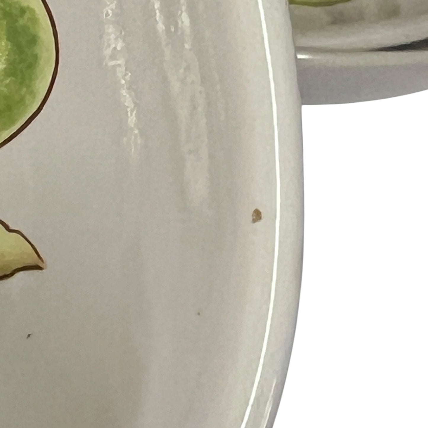 Close-up of a glossy off-white ceramic plate or dish with a small brown speck on the surface, showing part of a green painted motif at the left edge.