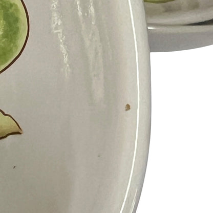 Close-up of a glossy off-white ceramic plate or dish with a small brown speck on the surface, showing part of a green painted motif at the left edge.