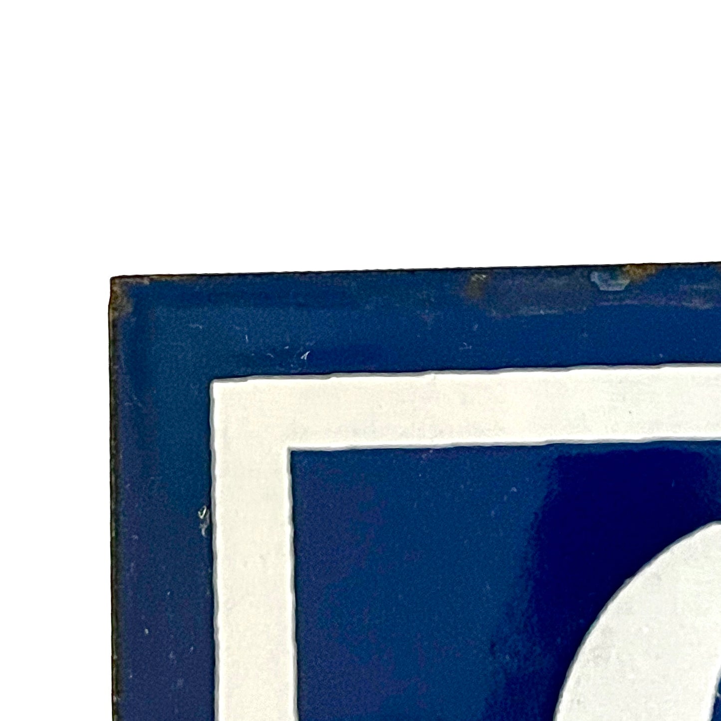 Close-up corner detail of a square decorative sign or tile with a dark blue field, white border, and part of a white curved shape visible against a white background.